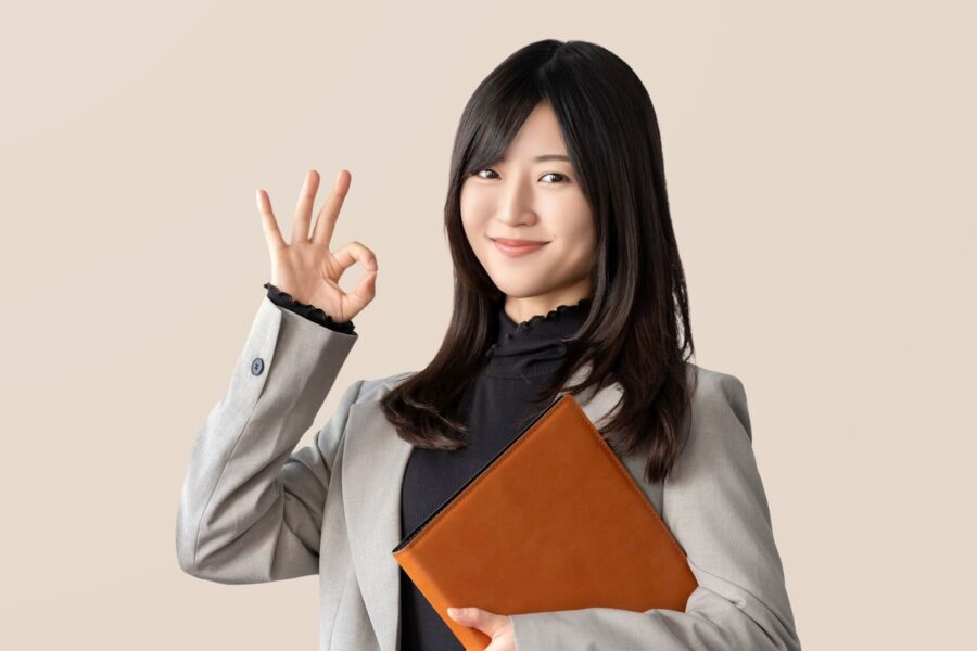 ビジネスカジュアル姿の女性が笑顔でOKサインを示し、手にビジネスファイルを持っている画像。バーチャルオフィスを利用することで得られるメリットやビジネス効果を象徴している。