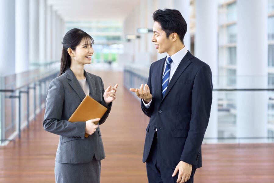 廊下で笑顔で会話するスーツ姿の男女ビジネスパーソン(信頼感・相談のしやすさをイメージ)